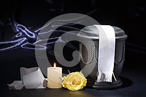 Black cemetery urn with burning candle and white ribbon yellow rose on deep blue background