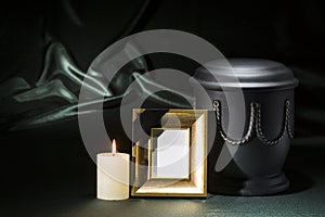 Black cemetery urn with golden mourning frame burning candle on deep green background