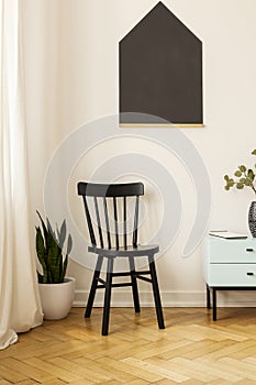 Black chair against white wall with poster in living room interior with cabinet. Real photo