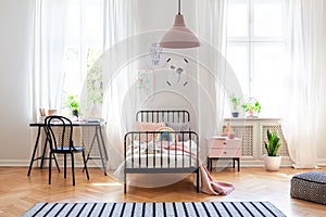 Black chair at desk next to bed in girl`s room interior with pink lamp above carpet