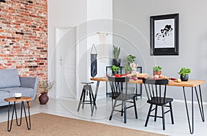 Black chairs at dining table and poster in flat interior with grey sofa against red brick wall