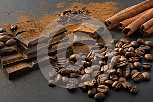 Black chocolate pieces, cinnamon and coffee beans, chocolate slabs spilled from grated chocolate powder on black dish. Close up.