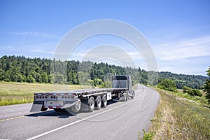 Black classic big rig semi truck tractor transporting empty flat bed semi trailer running on the summer winding highway road in