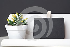 Black clip blackboard on modern pencil box and green plant in white pot on table and dark grey wall in office desk,mock up space