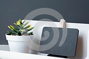 Black clip blackboard on modern pencil box and green plant in white pot on table and dark grey wall in office desk,mock up space