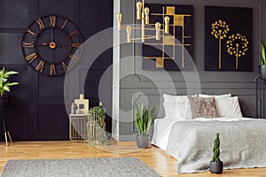 Black clock, golden chandelier, paintings and white bed in an elegant bedroom interior. Real photo