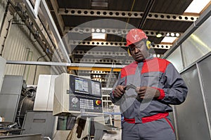 Black CNC Machine Operator In Protective Workwear Checking Measurements With Vernier Caliper Next To CNC Controller