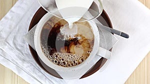 Black coffee in cup is poured with milk. Making caffeine drink. Close-up top view, wooden table, white napkin