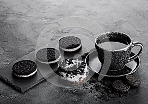 Black coffee cup with saucer and doughnut with black sandwich cookies on black stone kitchen table background. Space for text. Bre