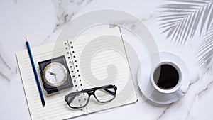 Black coffee in white ceramic cup, eyeglasses, vintage travelling leather case pocket watch,  pencil with note pad on white marble