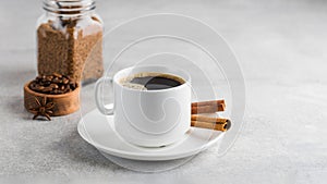 Black coffee in a white mug with cinnamon sticks on a saucer on a grey table. Copy space.