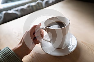 Black coffee in a white mug and saucer being held by a woman. The cuff of a cord jacket can be seen.