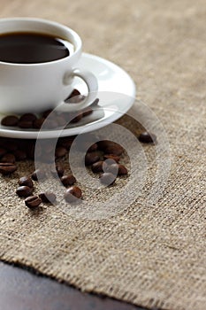 Black coffee in a white mug on a saucer and scattered coffee beans on burlap tablecloth