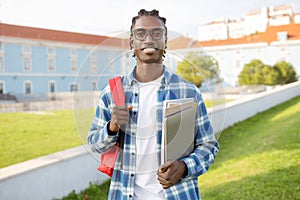 black college student guy holding workbooks, computer and backpack outdoor