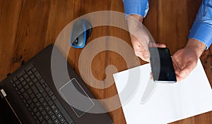 Black computer on old wooden desk. Mobile phone in man`s with blue shirt hand.