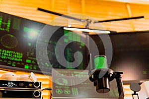 Black condenser microphone with green LED light, hacker coding interface on monitors, modern dark tech workspace setup