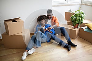 Black Couple With Digital Tablet Sitting Among Cardboard Boxes After Moving Home