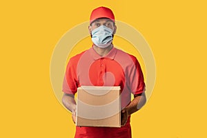 Black Courier Guy Delivering Cardboard Box Wearing Mask, Yellow Background photo