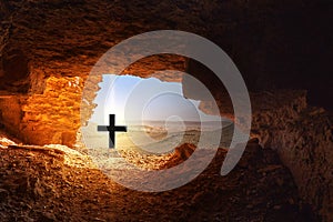 Black cross with bright light in front of dark cave in the dry hostile desert with boulders