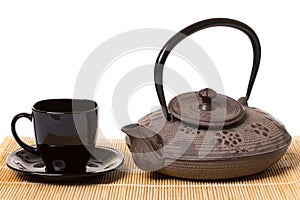 Black cup of tea on saucer and iron teapot on wooden mat.