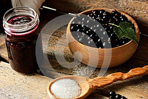 black currant berries in a wooden dish on a wooden table with a jar of jam, a wooden spoon with sugar, concatenation of