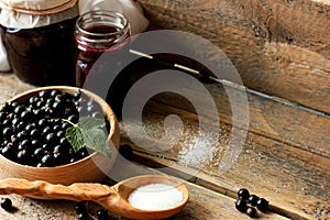 black currant berries in a wooden dish on a wooden table with a jar of jam, a wooden spoon with sugar, concatenation of