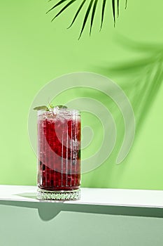 Black currant lemonade drink over green background. White table with sunlight and palm leaf hard shadow. Summer, tropical and