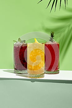 Black currant, orange and raspberry lemonade drink over green background. White table with sunlight and palm leaf hard shadow.