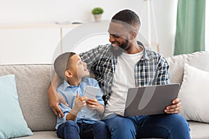 Black Daddy And Son Using Laptop And Phone At Home