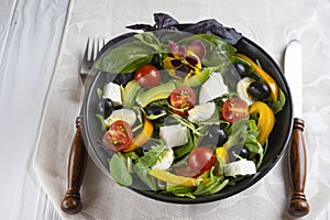 Black deep bowl of Greek salad, fork and knife on a white background.