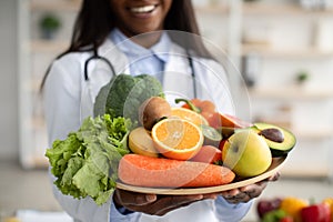 Black dietitian in lab coat holding bowl of fresh fruits and vegetables, recommending healthy plant based diet