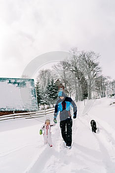Black dog follows a dad walking hand in hand with a little girl along a snowy road in the village