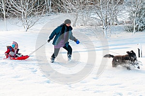 Black dog leading mother with daughter sledging behind through deep snow in a rural setting