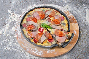 Black dough Pizza with meat slices, salami, ham, sliced tomatoes, mozzarella cheese served with fresh basil leaf on wooden plate