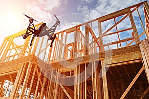 Black drone quadcopter with camera flying over residential construction home framing against a blue sky.