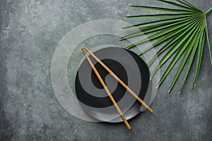 Black Empty Plate with Brown Bamboo Chopsticks Pal Tree Leaf on Dark Grey Stone Background. Asian Thai Chinese Cuisine Concept.