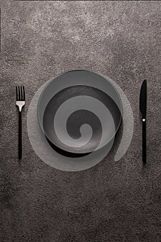 black empty plate and cutlery, fork and knife on a dark textured background. The concept of a layout for the design of a