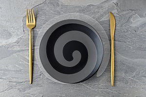 Black empty plate, matte golden knife and fork on grey slate background.