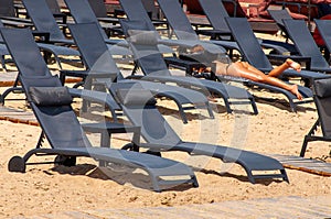 Black empty sun loungers on the sandy beach in the afternoon in rows, summer seaside holiday season, female tanned