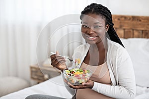 Black Expectant Woman Eating Fresh Vegetable Salad, Sitting On Bed At Home