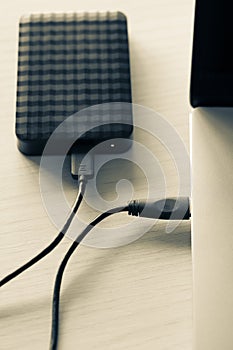 Black external hard disk, hdd on wooden table connected with mini usb cable to laptop, white background. Color image