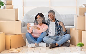 Black family chatting online with friends, showing key to new house on camera, sitting among carton boxes on moving day
