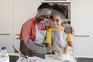 Black Father Baking With Daughter At Home, Checking Recipe On Digital Tablet