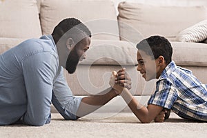 Black father competing in arm-wrestling with his little son at home