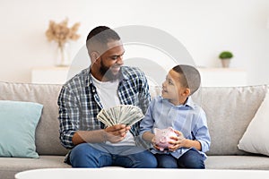 Black Father And His Son Putting Money In Piggybank Indoors