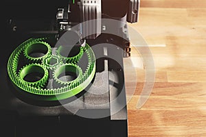 Black FDM-printer makes planetary gear machine part with visible infill from green filament in bright light.