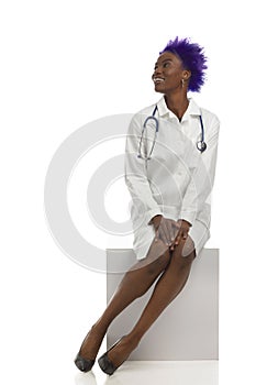 Black Female Doctor Sitting On White Box And Looking Up. Front View. Full Length