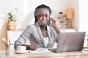 Black female entrepreneur talking on mobile phone, taking notes and smiling to camera