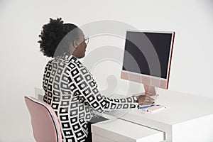 A black female office worker sitting at a white desk with desktop computer.