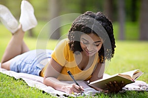 Black female student taking notes from textbook on picnic blanket in park